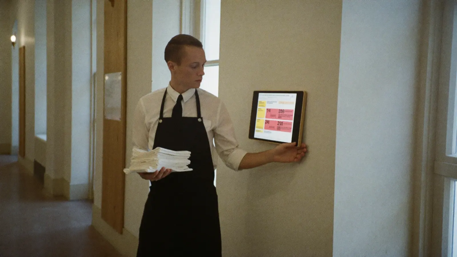 Een ober met een stapel servetten leest het weekrooster op een tablet aan de muur van een gang.