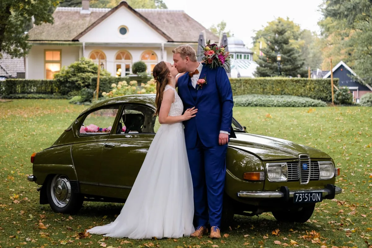 Een bruidspaar staat te zoenen naast een groene oldtimer in de tuin van de Agnietenberg