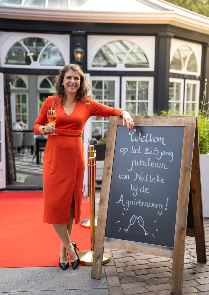 Nelleke Alleman-Dijkhuizen staat in een oranje jurk met een glas in haar hand naast een krijtbord waarop haar 12,5-jarig jubileum wordt aangekondigd