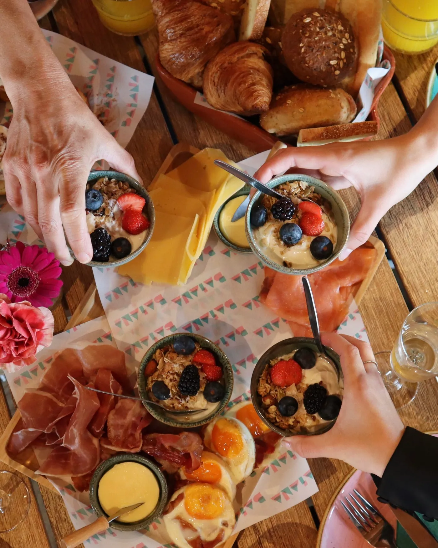 Een gedekte ontbijttafel van bovenaf, met croissants, yoghurt met fruit, bloemen en handen die een schaaltje pakken