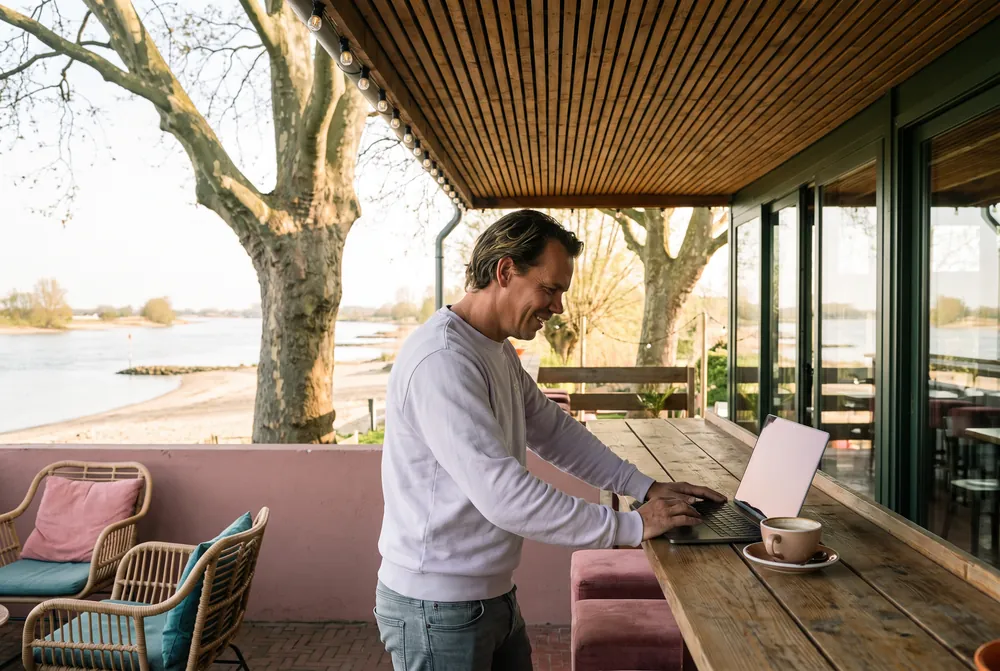 Mark Nijhuis staat aan de lange houten tafel op de veranda met een laptop voor zich, de Waal achter hem