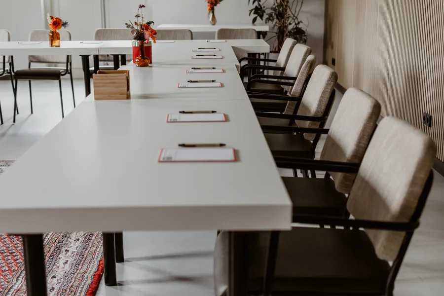 Een lange witte vergadertafel met notitieblokken en bloemen, onder houten balken