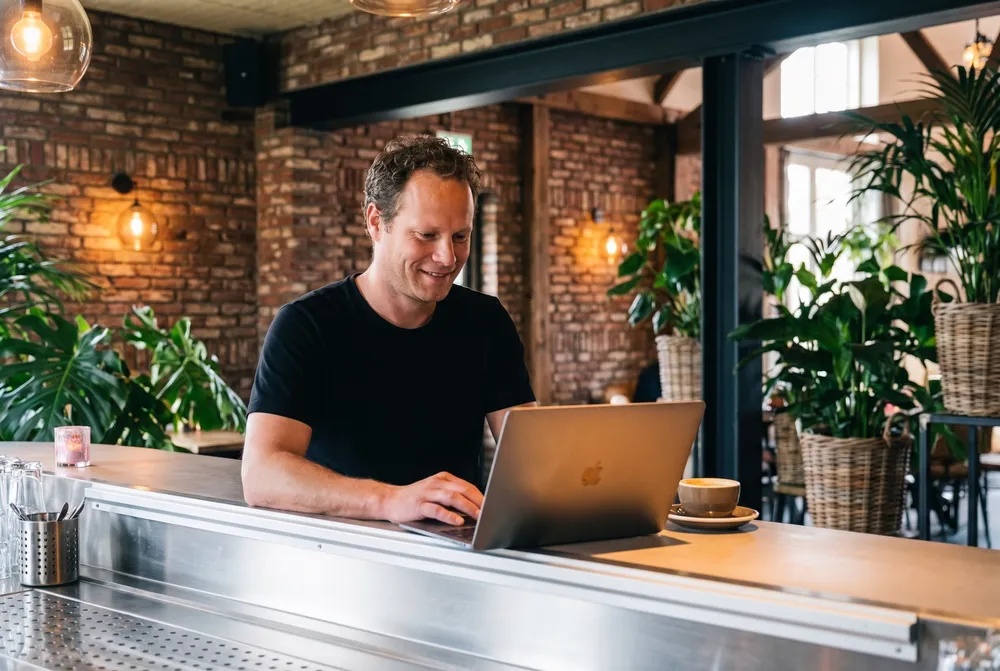 Pim Hamburg staat achter de bar van BOS & Co met een laptop voor zich, het restaurant met planten en bakstenen muren achter hem