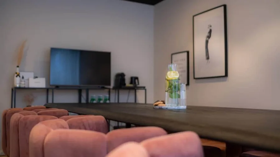 Een vergaderruimte met een lange tafel, roze stoelen en een groot scherm aan de wand