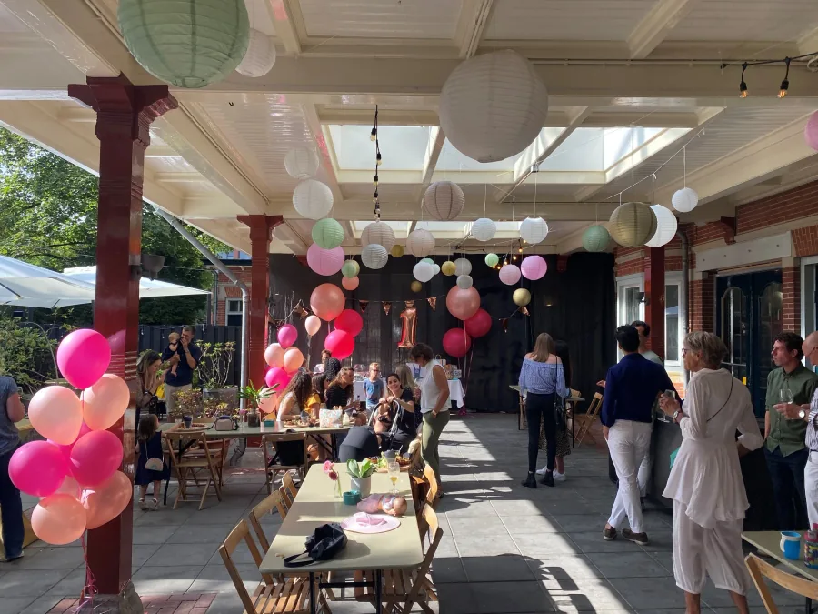 Gasten aan lange tafels in de tuin onder papieren lampionnen en ballonnen