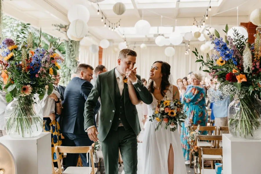 Een bruidspaar loopt tussen de gasten door naar binnen, met bloemen langs het pad